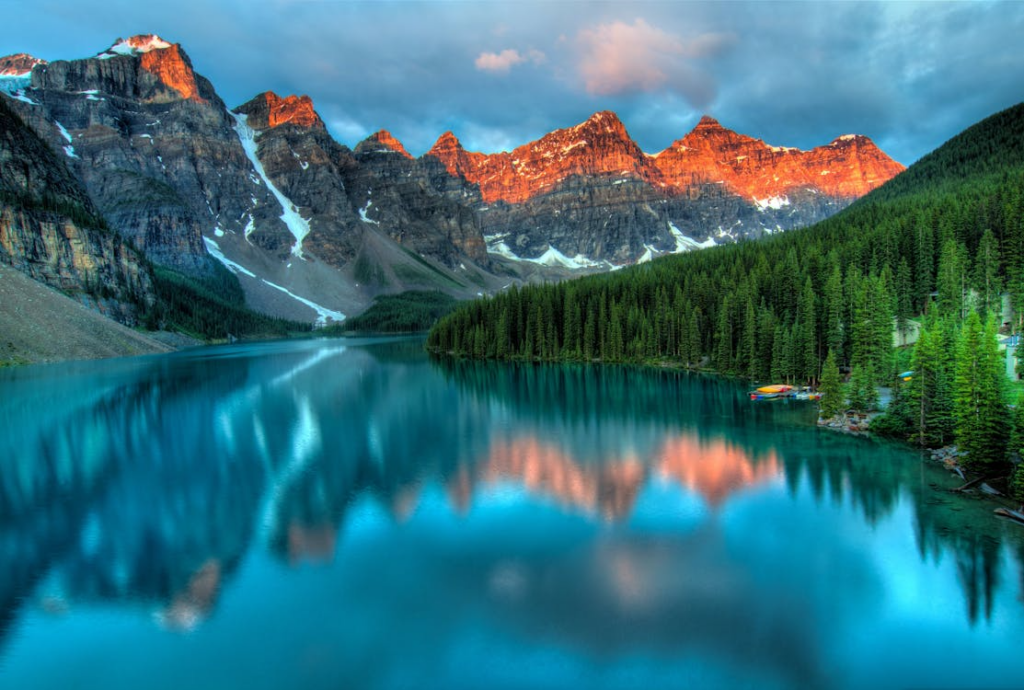 Beautiful rocky mountains tower over an alpine lake (Moraine Lake) at sunrise. The pink sunlight is just touching the peaks of the mountains. There is a forest on the lake shore.